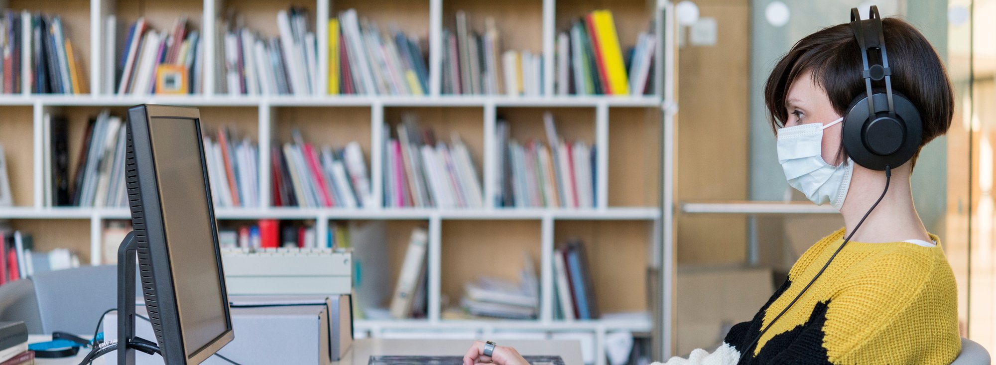 woman with facemask and headphones using a computer in a library